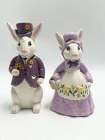 Photo of two ceramic Easter rabbits, male in purple jacket and top hat, female in purple dress with apron and bonnet, facing forward.