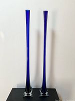 Front view of two tall cobalt blue glass vases with narrow midsection and flared tops standing side by side on a black surface against a white background.