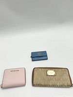 Three Michael Kors wallets: small blue wallet with MK logo, beige signature MK pattern wallet with plaque, light pink zip-around wallet