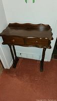 Front view of antique 2 drawer hall table against wall, showing drawer knobs and scalloped edges.