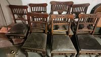 Photo shows the set of seven antique wooden chairs with carved backs and upholstered seats in a row, exhibiting wear and age-related patina.