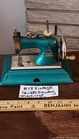 Front side view of antique child's sewing machine showing teal color, silver sewing area, hand crank, and part of the base.