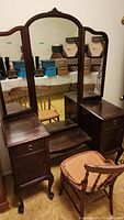 Full view of antique wooden vanity with triple mirrors and matching chair in front.