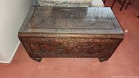 View of the full carved wooden chest coffee table topped with a rectangular glass panel on a carpeted floor.