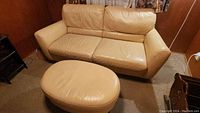 Cream leather loveseat and matching ottoman shown together in a room with brown walls and carpet. Loveseat shows signs of wear on armrests.