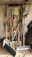 View of the full assortment of garden tools leaning against a pegboard wall, showing tools with wooden and metal handles including shovels, snow shovels, rakes, hoes, brooms, and hand tools.