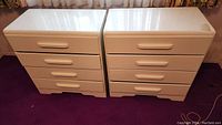 Pair of white wooden dressers each with 4 drawers, placed side by side on a purple carpeted floor. Shows minor wear on tops.
