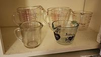 Five Pyrex measuring cups of various sizes arranged on a shelf, showing wear and one chipped cup.