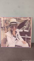 Front cover of Elton John Greatest Hits album showing Elton John sitting by piano in white suit, hat, and large glasses.
