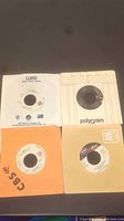 Four 7-inch records laid out showing labeled records and sleeves from different companies including CBS, Warner/Elektra/Atlantic, Polygram, and A&M. No cover art, sleeves only, records look worn.