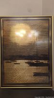 Framed brass etching artwork showing a waterfront scene with boats and a tall structure, under reflective glass.