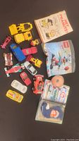 Overview of all items in lot on dark surface; shows toy vehicles, Peanuts book, two Ty Baby plush toys in packaging, Lego Classic set packaging