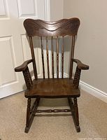 Front facing photo showing carved pressback, turned vertical spindles, broad armrests with turned supports, wooden flat seat, turned front legs and rockers.