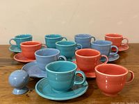 Overall display of a set of ten different colored Fiesta pottery cups and saucers plus one extra cup and a salt shaker on a wooden surface.