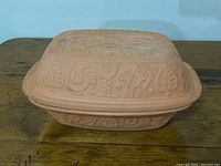 Side view showing the embossed animal and leaf motifs on the lid and side of the terracotta roasting pan.