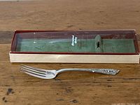 Fork with decorative Rosepoint pattern handle displayed outside original green velvet-lined box with clear plastic lid marked 'International Sterling'.