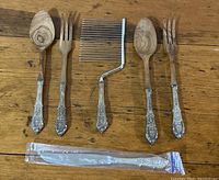 Top-down photo of five serving utensils showing detailed ornate Rosepoint sterling silver handles with wooden and metal working parts, plus one wrapped cake knife.