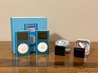 Two blue iPod Mini 6GB devices with original box and two iPod Nano 16GB (black and pink) with cords displayed on a wooden surface.