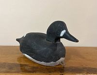 Front left side view showing the carved details and paint condition of the vintage wooden loon duck decoy, resting on a wooden surface against a plain wall.