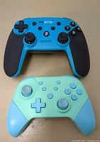Top view of two wireless game controllers, one black and blue, the other pastel green and blue, showing buttons and grips.