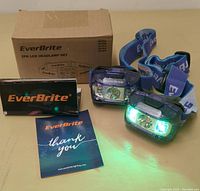 Two EverBrite LED headlamps with blue adjustable straps, glowing bright green light, positioned next to EverBrite box and printed materials.
