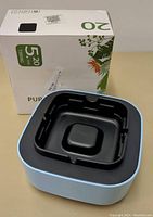 Photo of the air purification ashtray device showing square shape, black ashtray insert, and blue outer housing, placed next to the original box.