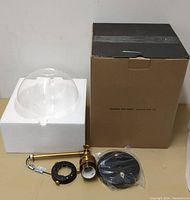 Clear glass dome shade on white foam packaging, brass mounting arm, black ceiling plate, and mounting ring on display beside cardboard box.