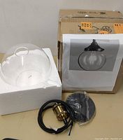 Photo of the ceiling light parts including clear glass globe, wired socket with black cord, and mounting plate, placed on styrofoam packaging with the box in background showing a printed product image.