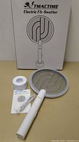 Complete product set including the TMACTIME electric fly swatter, circular charging stand, USB charging cable, and instruction manual on a white background near the product box.