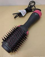 Electric hair dryer brush displayed on plain surface showing black and pink design, soft bristles with pink tips, power cord wrapped with velcro strap.