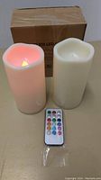 Two ivory flameless LED candles and remote control, one candle lit with warm yellow and red flame effect, cardboard box in back