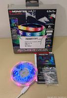 Photo shows the Monster Smart Illuminesce RGB+IC LED Light Strip rolled up and lit with vibrant colors alongside its packaging which shows some damage.