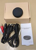Photo showing the mini Bluetooth music receiver device with associated RCA cables, USB power cable, 3.5mm to 1/4 inch adapter, a cardboard box, and instruction manual for user guidance.