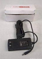 OMNIHIL switching adapter with power cable coiled neatly, adapter specifications label visible on black plastic housing, shown with packaging box in background.