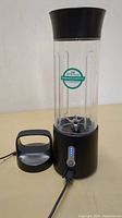 Front view of the portable electric juicer cup, showing clear BPA-free marked cup with blade assembly inside, black base with power button and LED battery indicator lights, connected to charging cable, and detached black lid placed beside it.