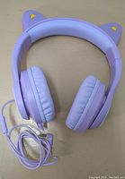 Light purple over-ear headphones with cat ear design, viewed from front on beige surface. Shows headband and padded ear cups with wired cable coiled.