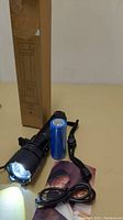 Photo of the black rechargeable flashlight with blue battery, USB cable, wrist strap, and cardboard packaging in the background with the light switched on.