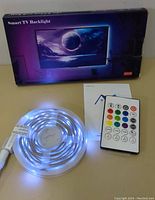 Photo of LED strip light coiled on spool, white light active, alongside remote control and instruction manual, with packaging in background.