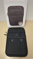 Photo showing the black universal tablet sleeve in front of its packaging. Sleeve has a zipper and multiple front pockets labeled with icons for organizing a phone and cables.
