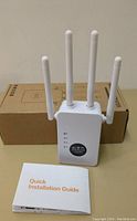 Front view of wireless repeater device with four external antennas standing upright on surface, with quick installation guide booklet in front and cardboard box behind.