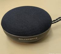 Front angled view of compact round wireless Bluetooth speaker covered in black mesh fabric with 'Soundperts' branding on the side.