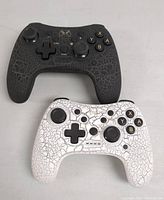 Top view of two wireless gaming controllers, one black and one white, both with crackle surface design and labeled buttons.