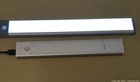 Two rechargeable sensor motion lights turned on, revealed rectangular shape with LED light panels. One light is longer with gray sensor end and the other is shorter with white body and pink button.