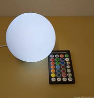 White ball-shaped lamp humidifier glowing with light next to a remote control with color and timer buttons, placed on a yellow surface.