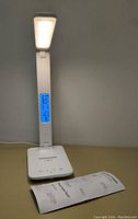 Front view of white digital desk lamp turned on showing blue-lit digital display with time and temperature. Instruction sheet placed beside the lamp.