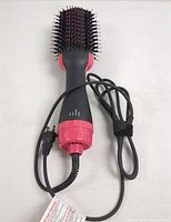 Front view of hot air brush blower with pink and black paddle brush head, power cord coiled beside it.