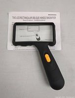 Magnifying glass with rectangular lens and black ergonomic handle with yellow LED switch, lying on instruction sheet detailing specifications and features.