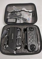 Open carrying case showing mini drone and remote control securely stored in foam cutouts. Accessories such as cables and manuals visible in mesh pouch in case lid.