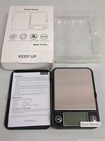 Open scale showing stainless steel platform, digital display with unit and on/off buttons, user manual, and clear plastic cover.