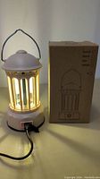 Photo of the USB rechargeable lantern lamp powered on with glowing warm light. Shows hanging handle, vertical bars around the light source, power switch, and USB port with cable plugged in.